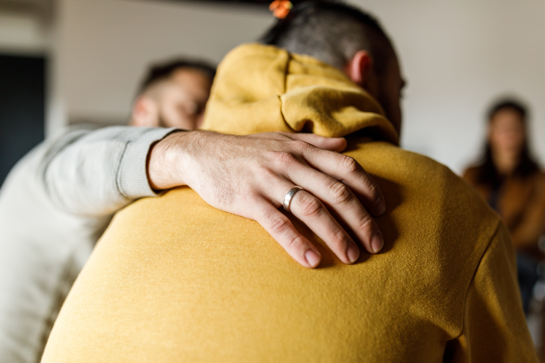 The back of a man with a yellow sweater. Another hand is placed on the back signaling care.