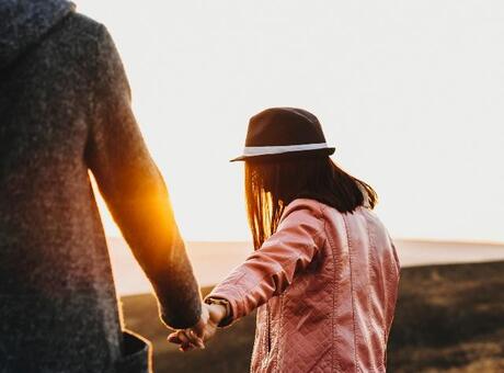 Person in a pink jacket and hat walking across an open landscape while holding another person’s hand.  Warm sunset light shines between them, creating a bright glow against the natural background.