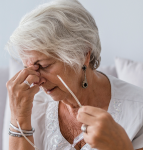 An older woman with gray hair taking off her glasses and holding her nose grimacing.