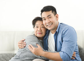 A man and an older woman hugging each other on a couch while smiling.