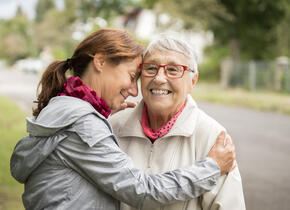 A middle aged woman hugging an older woman. Both are outside and smiling.